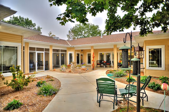 Outdoor courtyard area of a senior living facility with a paved walkway, garden beds with plants, bird feeders, and green chairs arranged for seating. The building surrounds the courtyard with large windows and a covered patio area with tables and chairs.