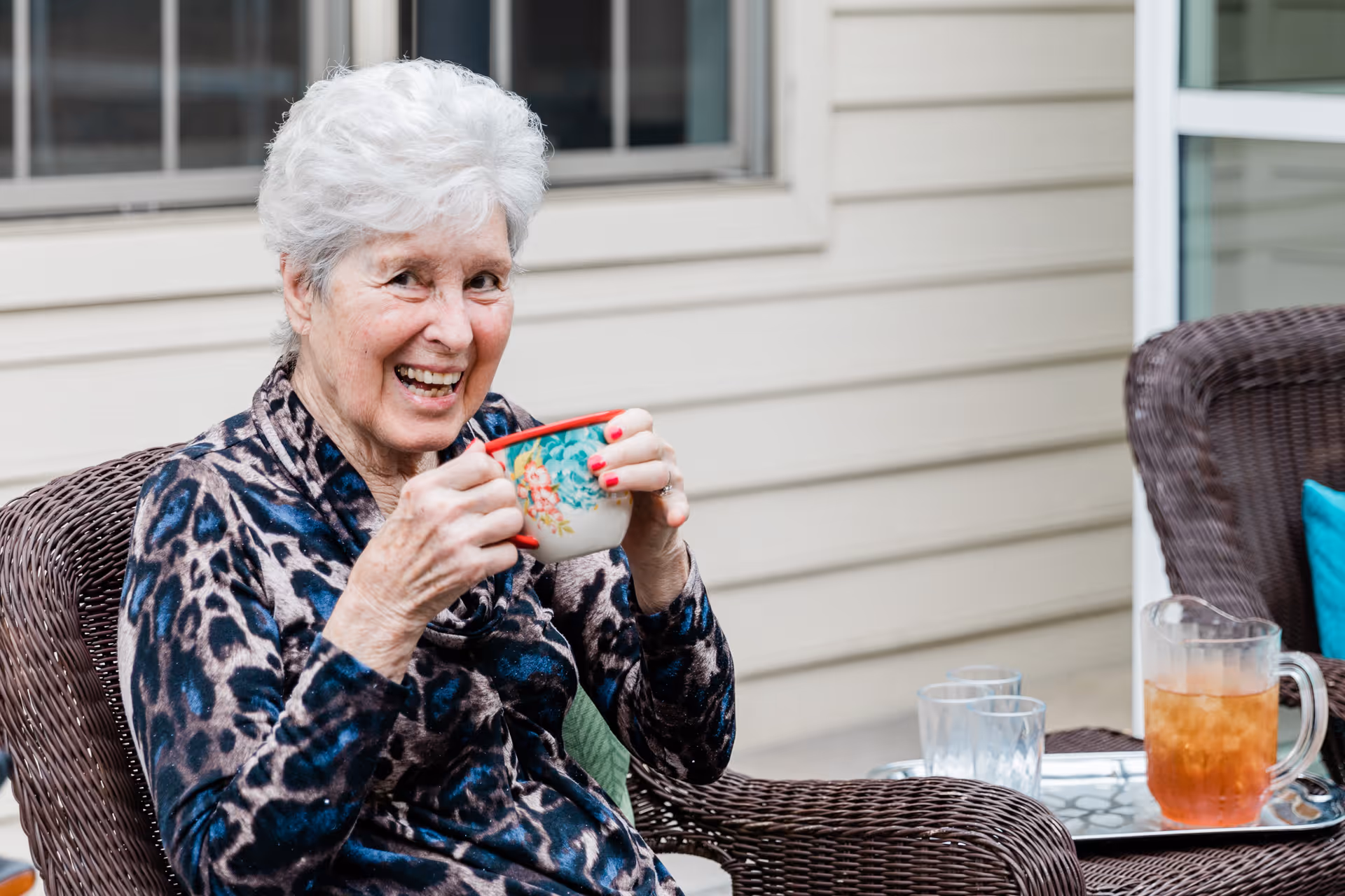 An elderly woman with white hair sitting outdoors on a wicker chair, smiling and holding a floral-patterned cup. There is a tray with a pitcher of iced tea and two glasses on a table beside her. The background shows the exterior siding of a building and a window.