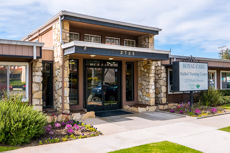 Exterior front entrance of Royal Care Skilled Nursing Center building with stone and brown paneling, glass double doors, and a sign displaying the facility name and address. There are flower beds and greenery around the entrance.