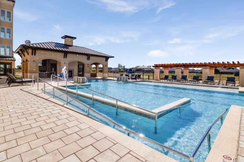 Outdoor swimming pool with a handicap ramp, lounge chairs, and a covered pavilion under a blue sky.