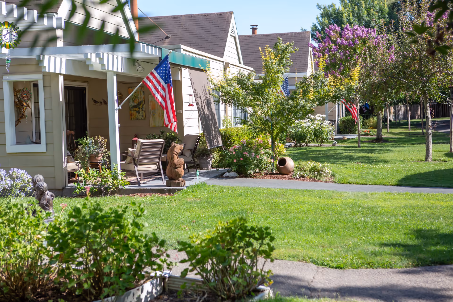 A peaceful outdoor scene at O'Connor Woods featuring a row of single-story residential units with porches. Each porch is decorated with chairs, plants, and American flags. The area is surrounded by well-maintained green lawns, trees, and flowering bushes under a clear blue sky.