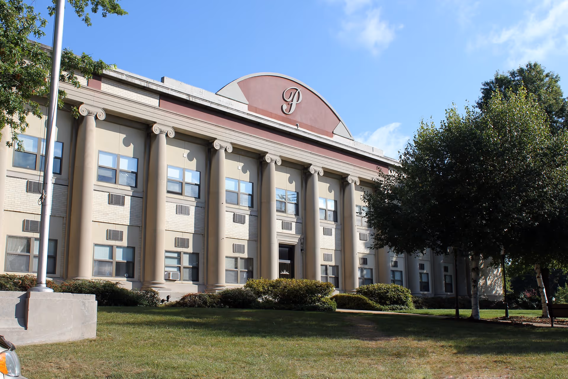 Exterior view of a large, classical-style building with tall columns and multiple windows, surrounded by green grass and trees under a blue sky.