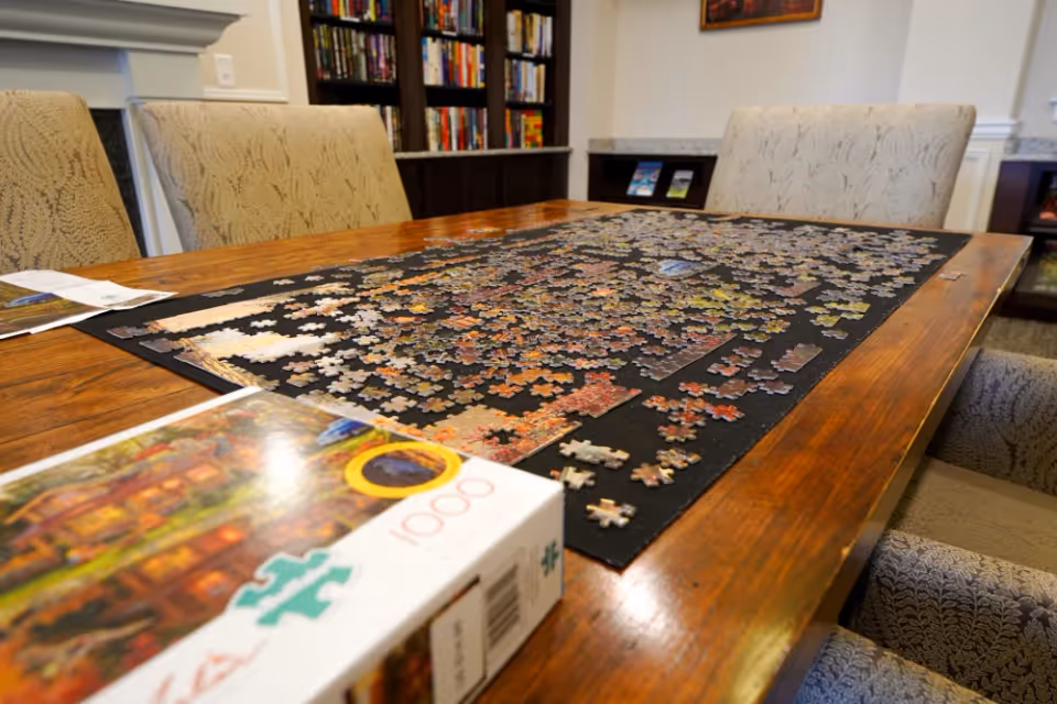 A wooden table with a partially completed jigsaw puzzle on a black puzzle mat. The puzzle box is visible in the foreground. Surrounding the table are upholstered chairs with a patterned fabric. In the background, there is a bookshelf filled with books and a countertop with some brochures or pamphlets.