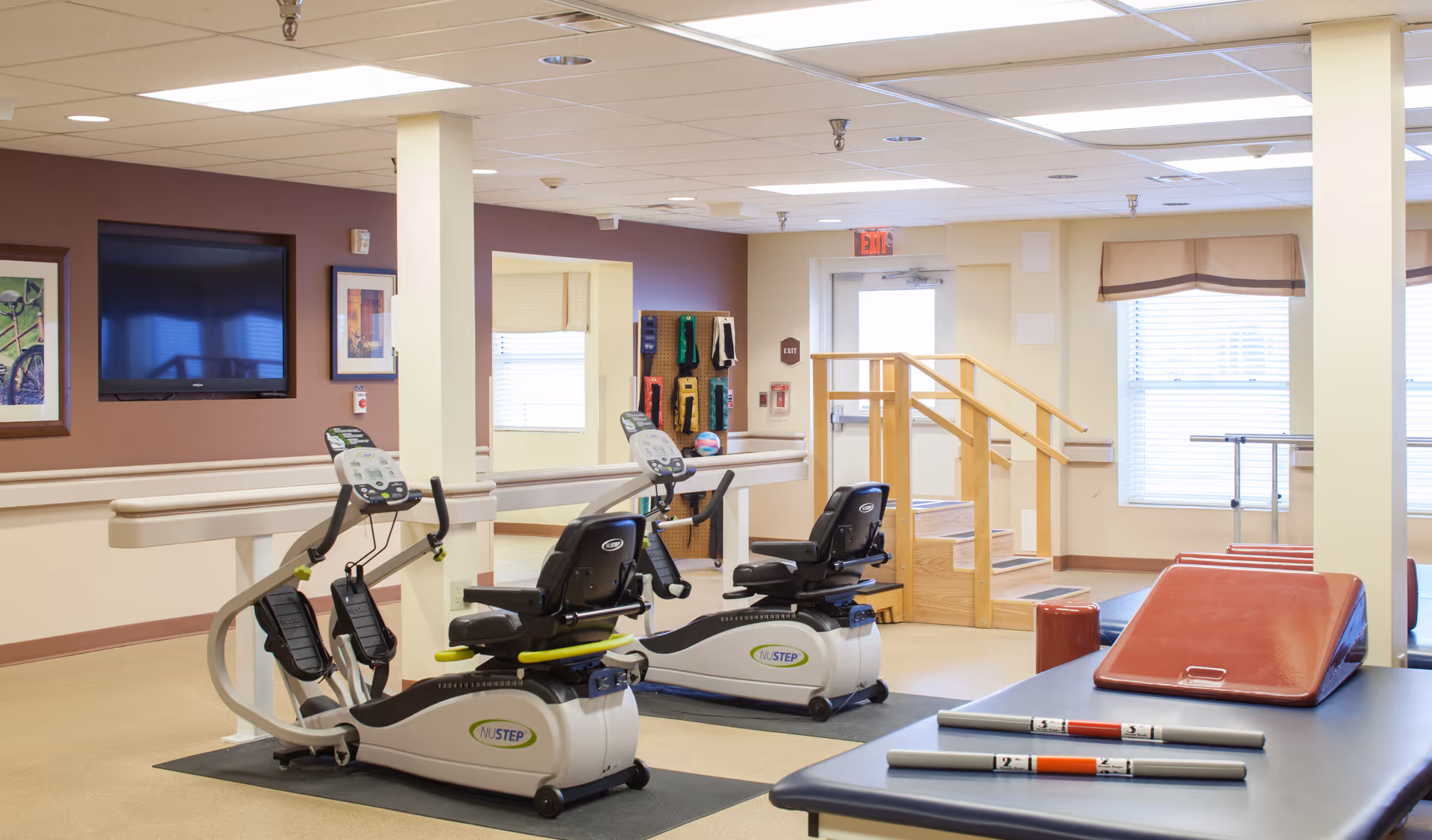 A well-lit exercise room in a senior living facility featuring two NuStep recumbent cross trainers, a set of stairs with handrails, resistance bands hanging on the wall, a large flat-screen TV, framed pictures, and exercise mats with weighted bars on a table.