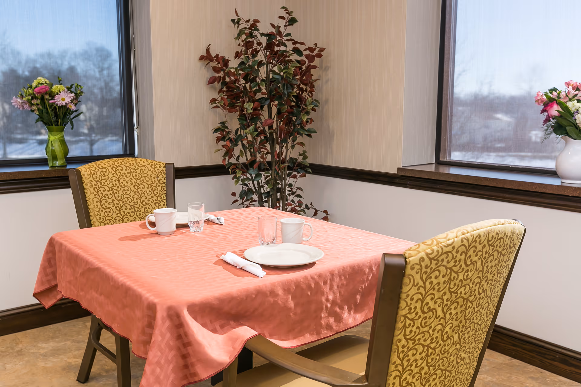 A small dining area with a square table covered by a pink tablecloth, set with two white plates, two white mugs, two glasses, and two napkins. Two upholstered chairs with yellow patterned fabric are positioned at the table. There are two windows with a view of a snowy landscape outside, and two vases with flowers on the window sills. A tall potted plant is placed in the corner between the windows.