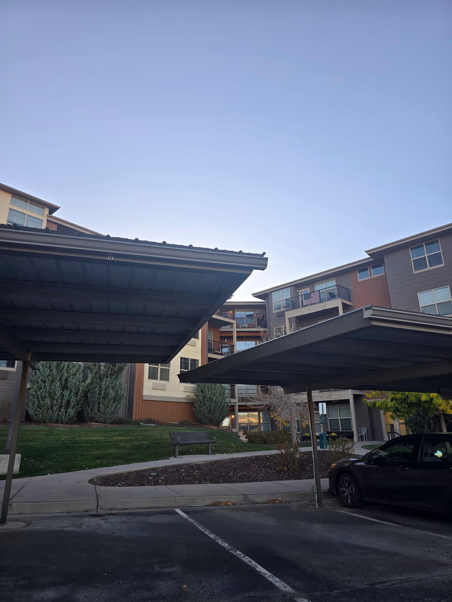 Covered parking spaces with a black car parked in one of them, in front of a multi-story residential building with balconies and windows. There is a small grassy area with bushes and a bench near the building, under a clear sky.