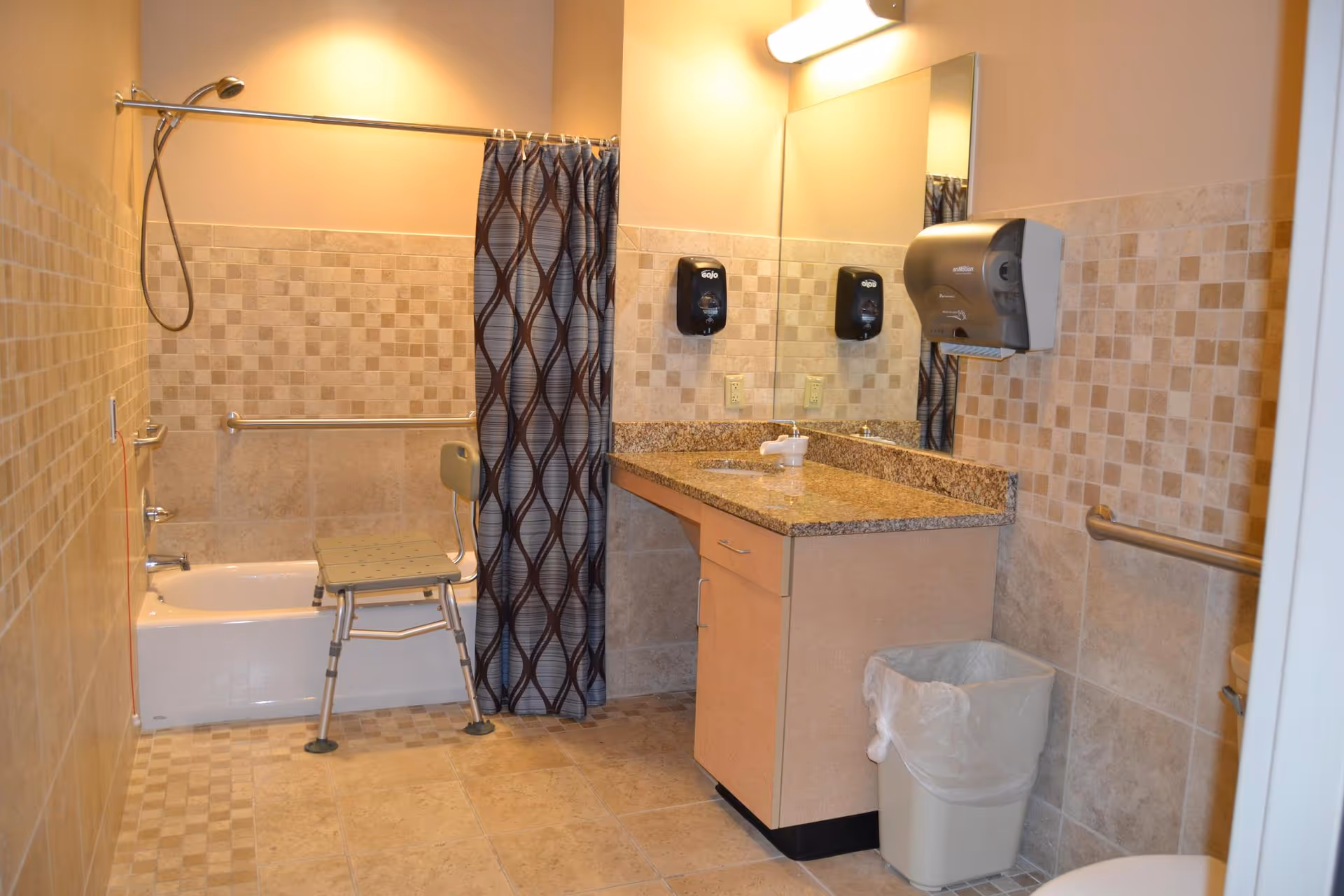 A bathroom with beige tiled walls and floor, featuring a bathtub with a showerhead and a shower chair inside. There is a shower curtain with a geometric pattern. A granite countertop with a sink, soap dispenser, and paper towel dispenser is visible, along with a trash can and grab bars on the walls for accessibility.