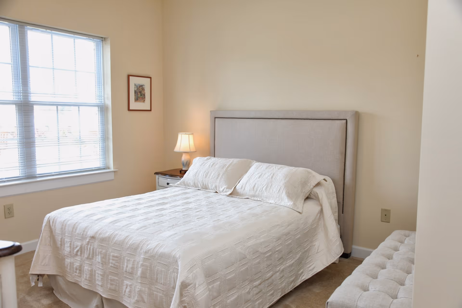 A bedroom with a neatly made bed featuring a light-colored headboard and white bedding. There is a nightstand with a lamp on the left side of the bed, a window with blinds letting in natural light, a small framed picture on the wall, and a cushioned bench at the foot of the bed.