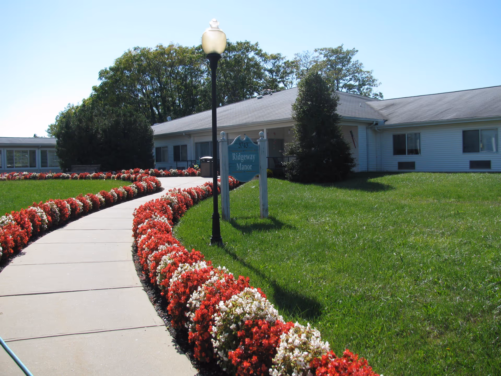A curved concrete walkway bordered by red and white flowers leads to a single-story white building with a gray roof. There is a lamppost and a sign that reads '5743 Ridgeway Manor' near the walkway. Green grass and trees surround the area under a clear blue sky.