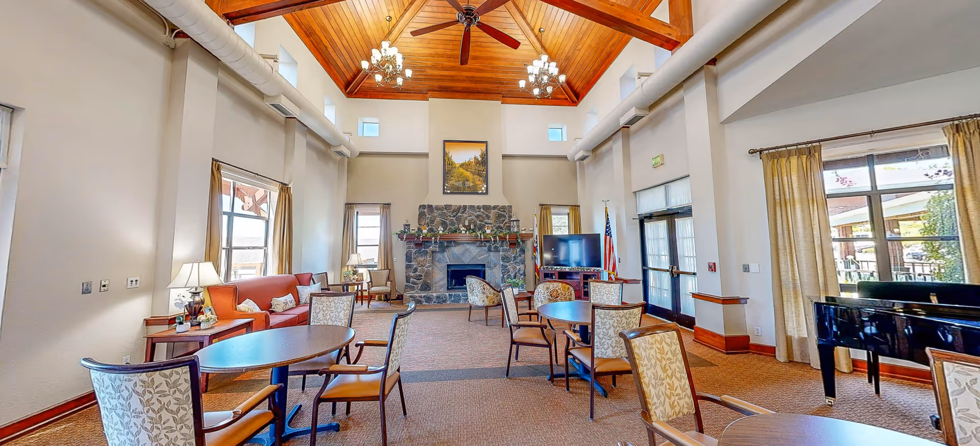 A spacious senior living facility common area with high wooden ceilings, ceiling fans, and chandeliers. The room features multiple round tables with chairs, a red sofa, armchairs, a stone fireplace, a flat-screen TV, and large windows with curtains letting in natural light. There is also a black grand piano near the windows.