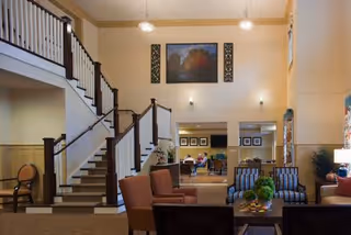 Interior view of a senior living facility lounge area with comfortable seating including armchairs and sofas arranged around a coffee table. A staircase with dark wooden railings leads to an upper floor. The walls are decorated with framed artwork and sconces, and pendant lights hang from the ceiling. In the background, there is a dining area with people seated at tables.