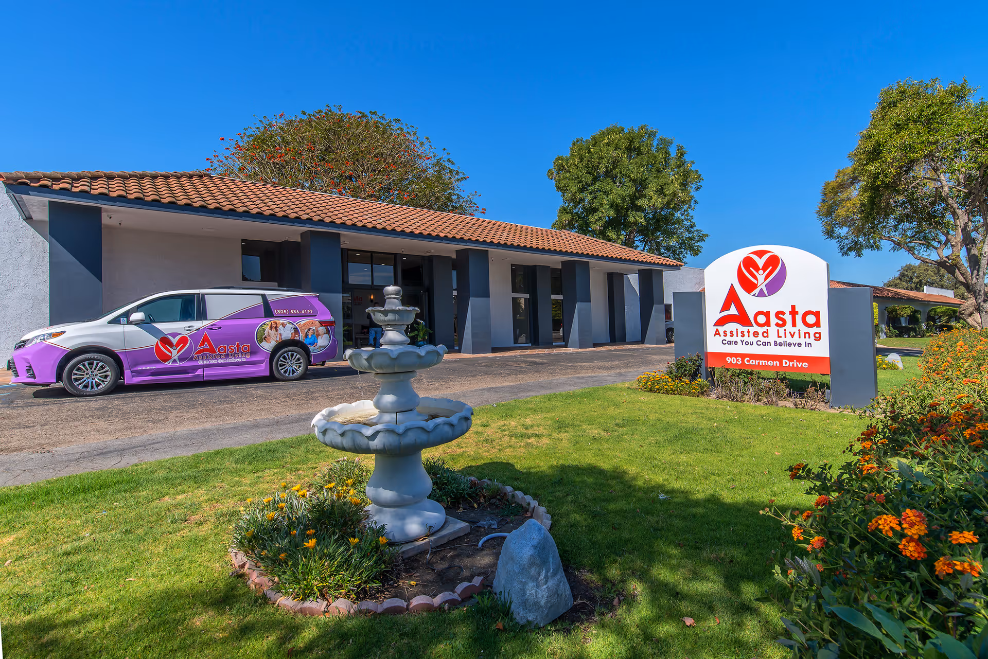 Exterior view of Aasta Assisted Living of Camarillo building with a tiled roof, a decorative fountain in the foreground, a purple van with the facility's logo parked in front, and a large sign displaying the facility's name and address on a green lawn with trees and flowers.