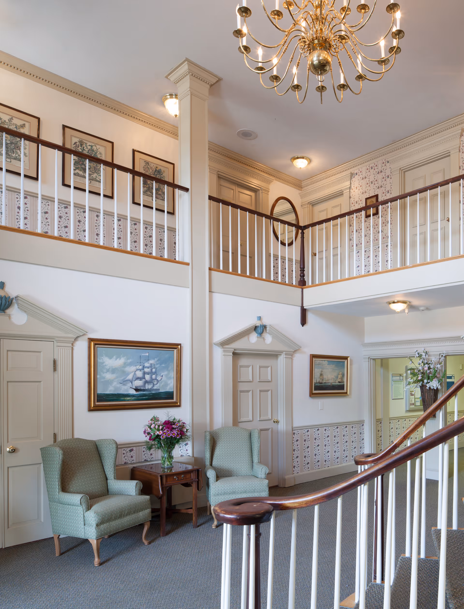 Interior view of a senior living facility lobby or common area with two green upholstered armchairs and a wooden side table with a vase of flowers. The space features a staircase with a wooden handrail, white spindles, and a second-floor balcony with white railings. The walls are decorated with framed paintings of ships and floral wallpaper below the chair rail. A large gold chandelier hangs from the ceiling.