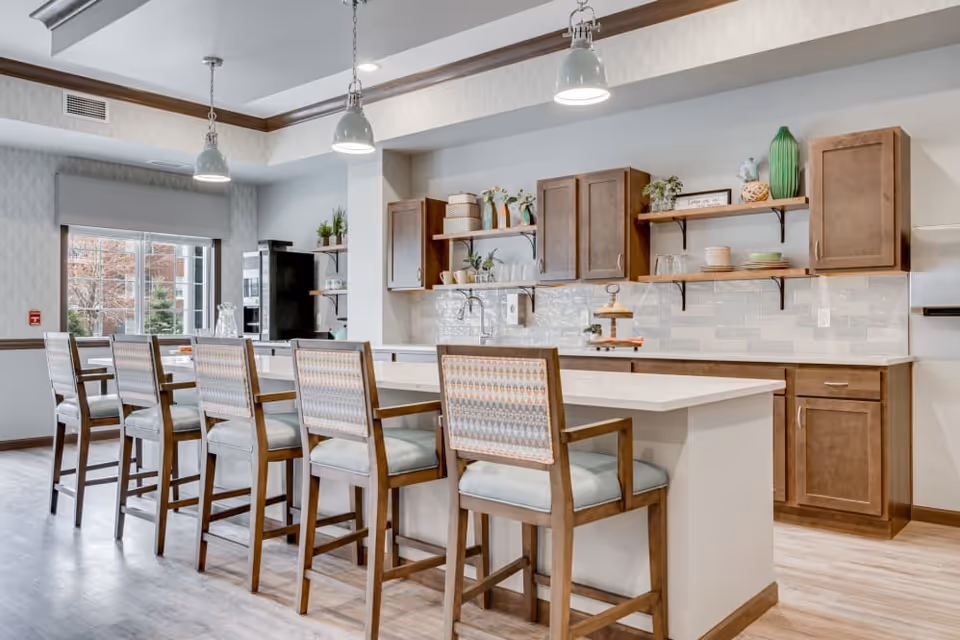 A modern kitchen area with a long white island countertop surrounded by six wooden chairs with patterned cushions. The kitchen features wooden cabinets, open shelves with decorative items and dishes, a refrigerator, and three pendant lights hanging from the ceiling. A large window allows natural light to brighten the space.