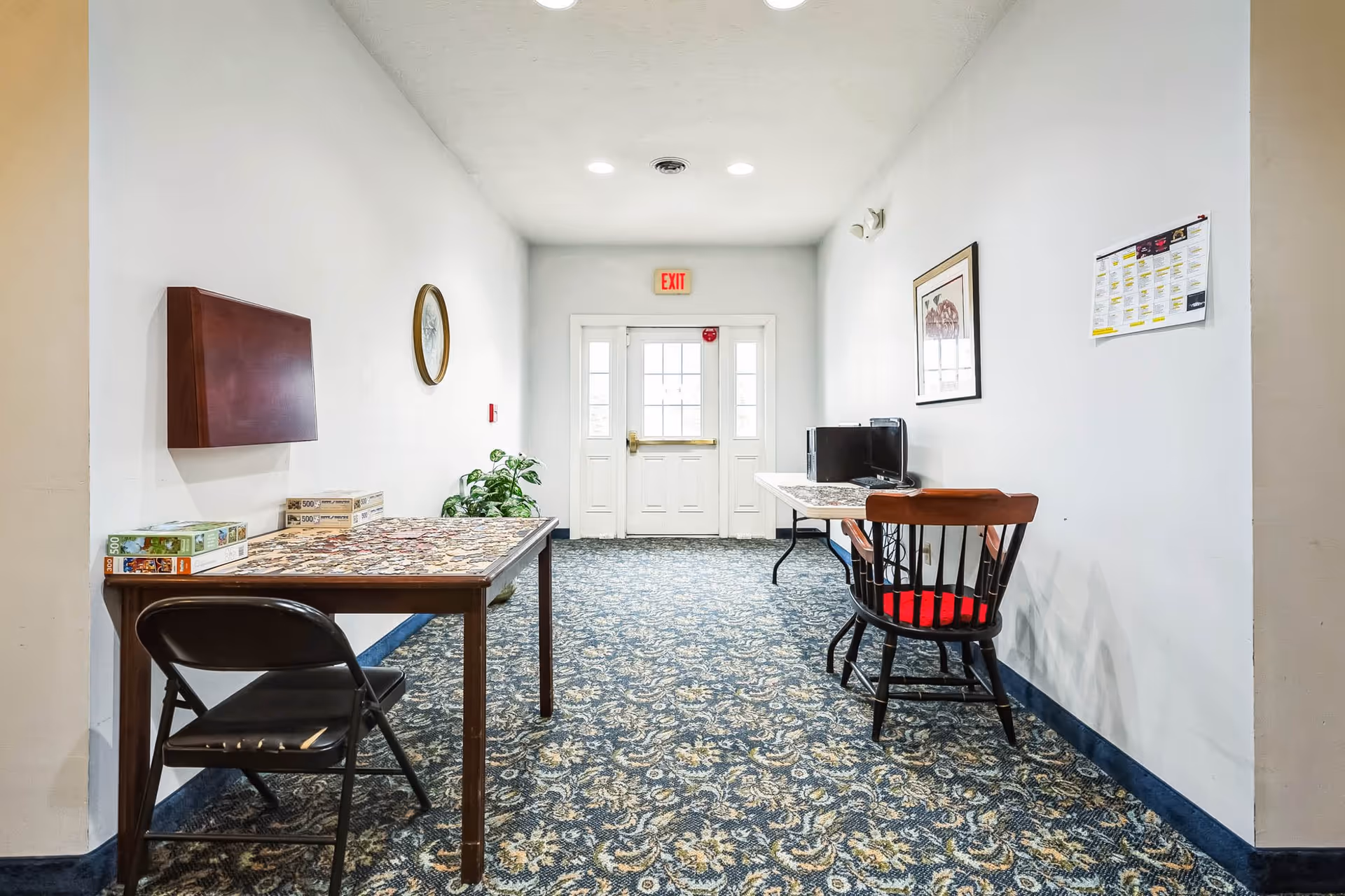 A hallway with patterned carpet and white walls featuring two tables along the sides. One table has a partially completed jigsaw puzzle and puzzle boxes with a black folding chair, while the other table has a computer monitor and a wooden chair with a red cushion. There is a clock on the left wall, a framed picture and a calendar on the right wall, and a white exit door with windows at the end of the hallway.
