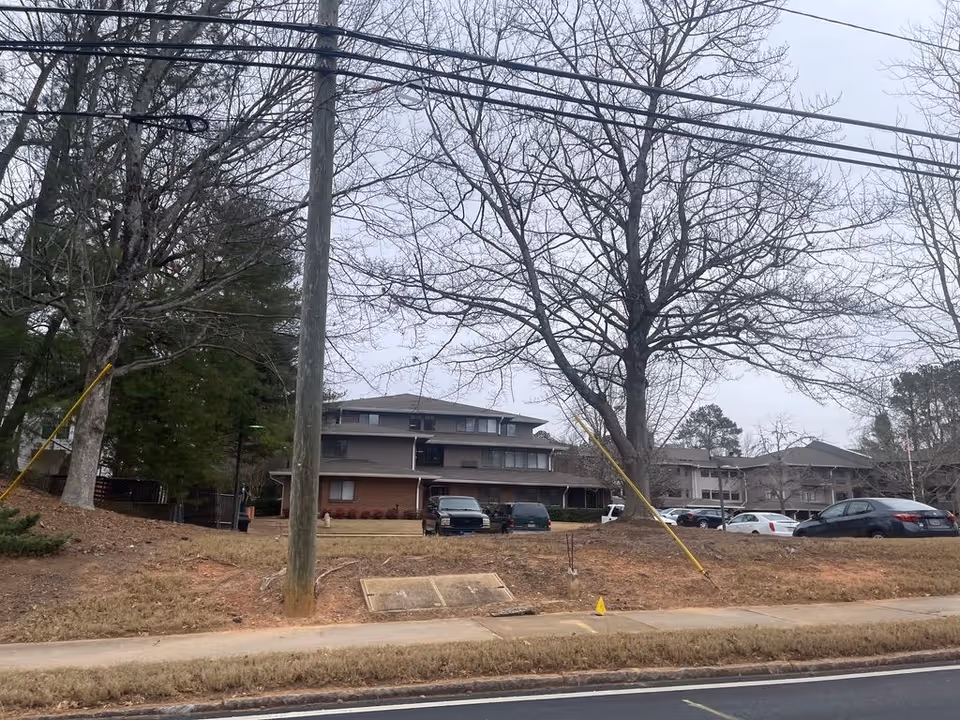 Front exterior of a multi-story senior living building with parked cars, leafless trees, and power lines in the foreground.