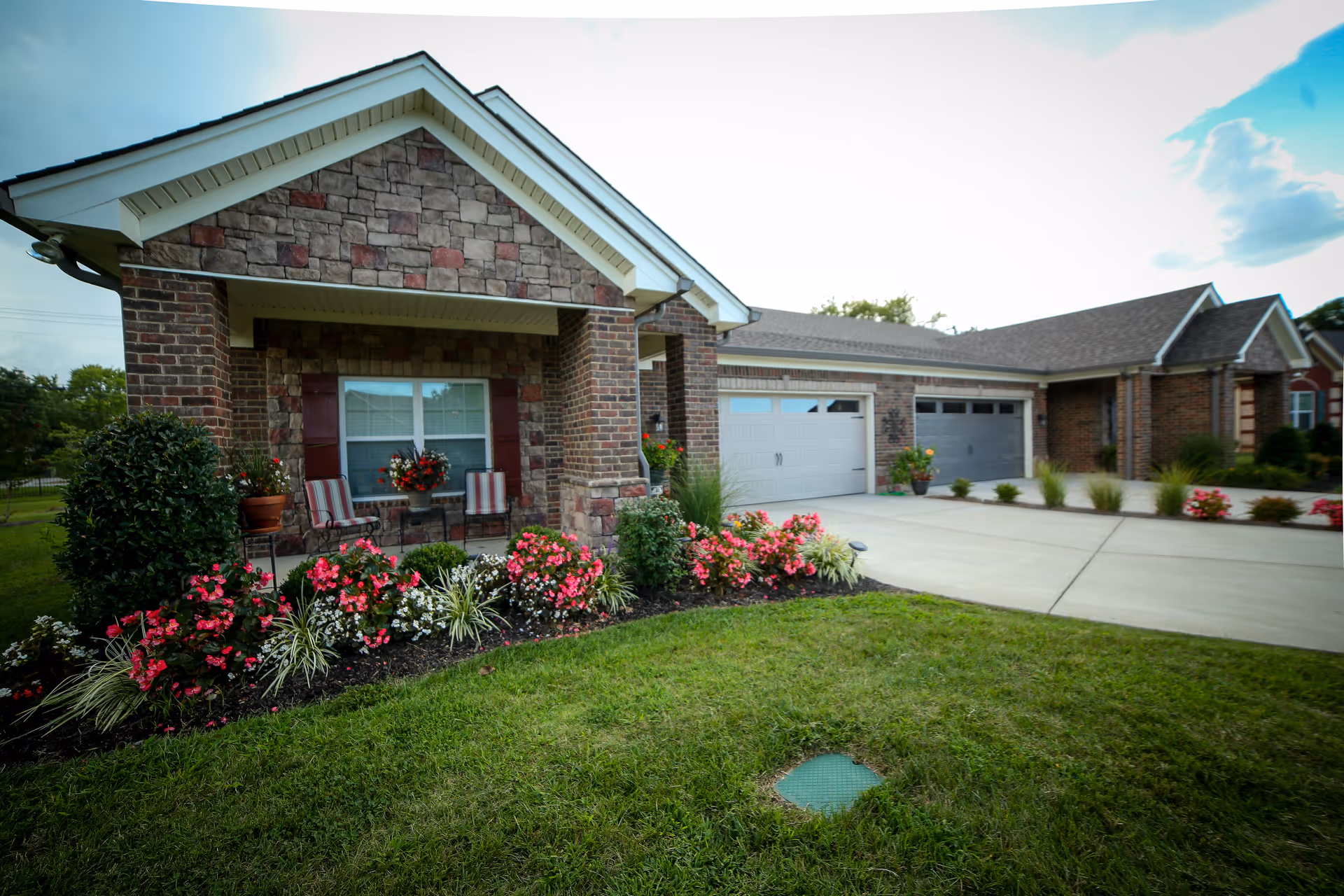 Front exterior view of a single-story brick and stone senior living facility with a covered porch featuring two chairs and a small table with flowers. The driveway leads to two garage doors, and there are well-maintained flower beds with pink and white flowers along the front of the building.