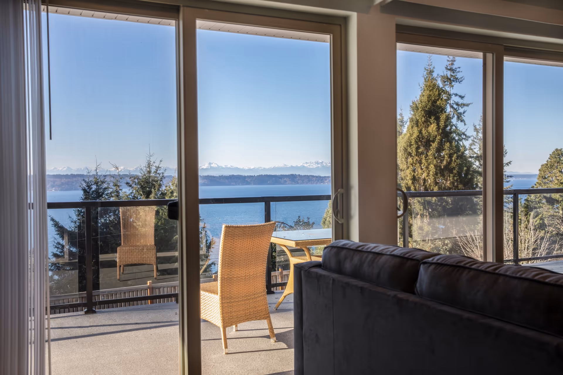 Living room with a sofa facing sliding glass doors that open to a balcony with wicker chairs and a water and mountain view.