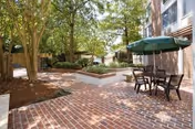 Outdoor patio area with brick flooring, raised garden beds, trees providing shade, and a table with four chairs under a green umbrella next to a building.