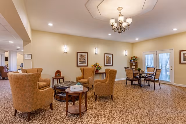 A cozy common area in a senior living facility with patterned carpet, four upholstered armchairs arranged around a round ottoman, a small side table with a tissue box, a dining table with four chairs near glass doors, framed artwork on the walls, and a chandelier hanging from the ceiling.