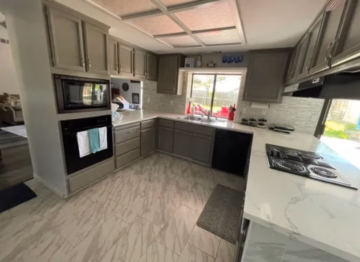 Modern kitchen with gray cabinets, white marble countertops, a built-in microwave and oven, electric stovetop, dishwasher, and a window above the sink overlooking a backyard with red outdoor furniture.