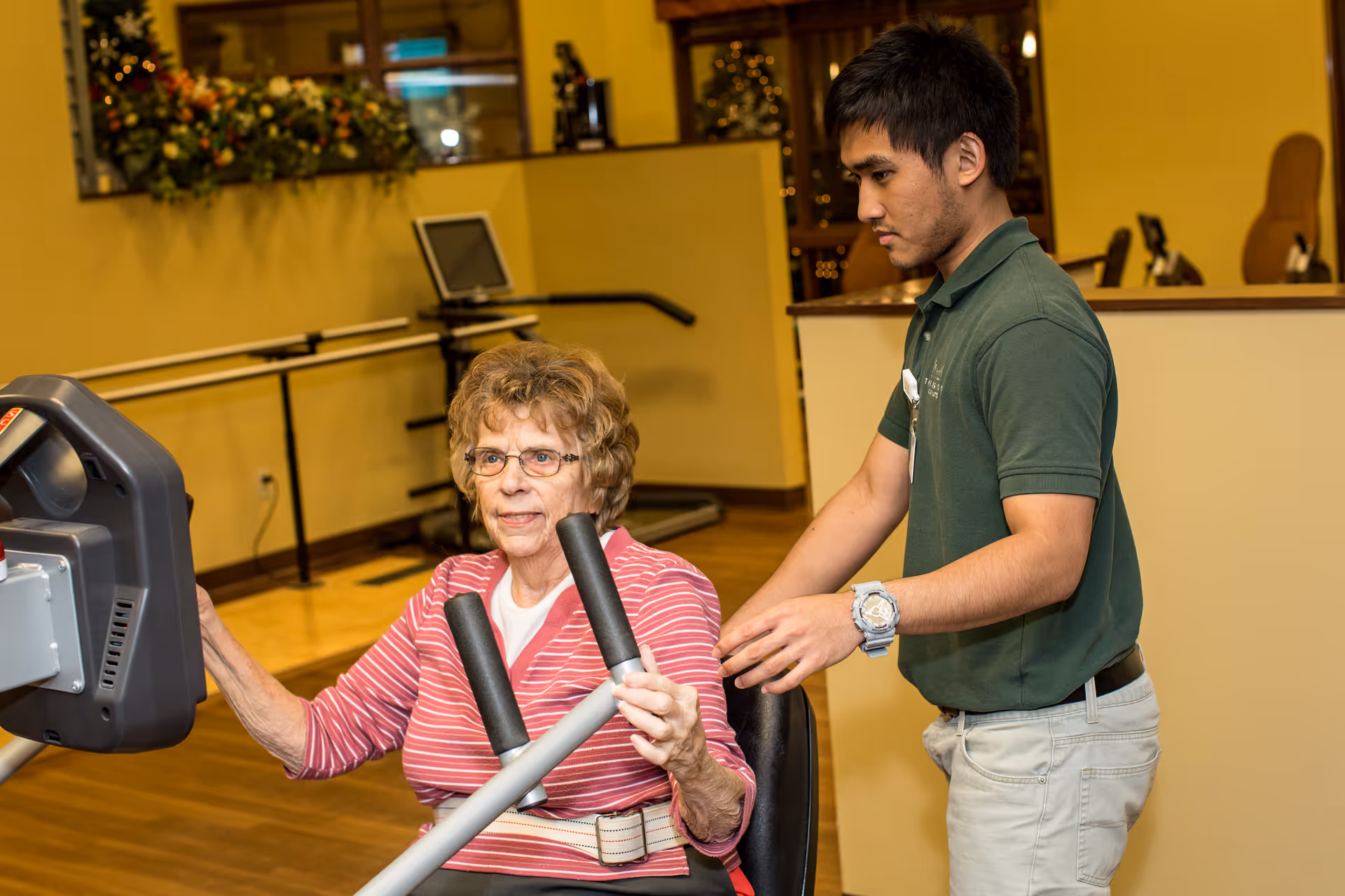 An elderly woman in a pink striped shirt is using a seated exercise machine in a fitness room, assisted by a young male staff member wearing a green polo shirt and beige pants. The room has wooden floors and yellow walls with exercise equipment and a mirror in the background.