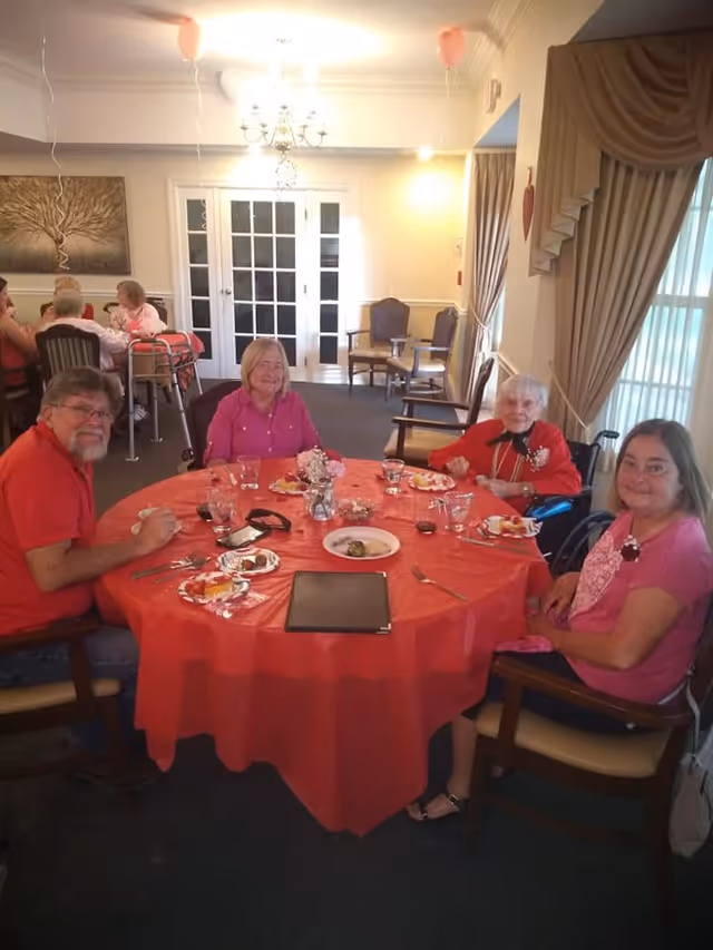 Four elderly people sitting around a round table covered with a red tablecloth in a dining room. They are smiling and appear to be enjoying a meal together. The room has beige walls, a chandelier, and large windows with curtains. There are additional people and chairs visible in the background.