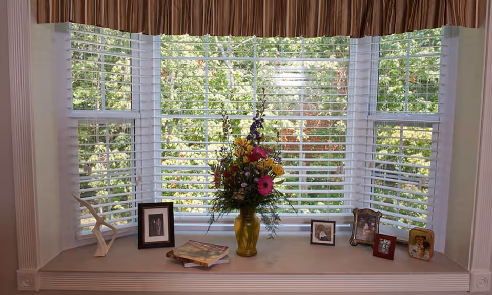 A window with white blinds partially open, showing green trees outside. On the windowsill, there is a yellow vase with a colorful bouquet of flowers, several framed photos, a small sculpture, and a couple of books.
