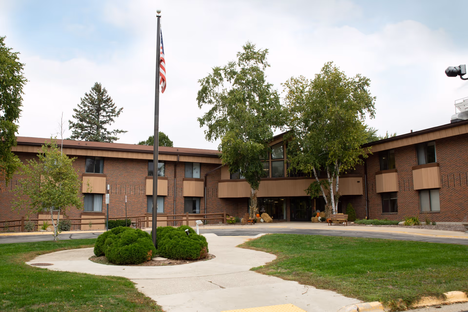 Two-story brick senior living facility entrance with an American flag on a flagpole, trees, and a circular driveway.