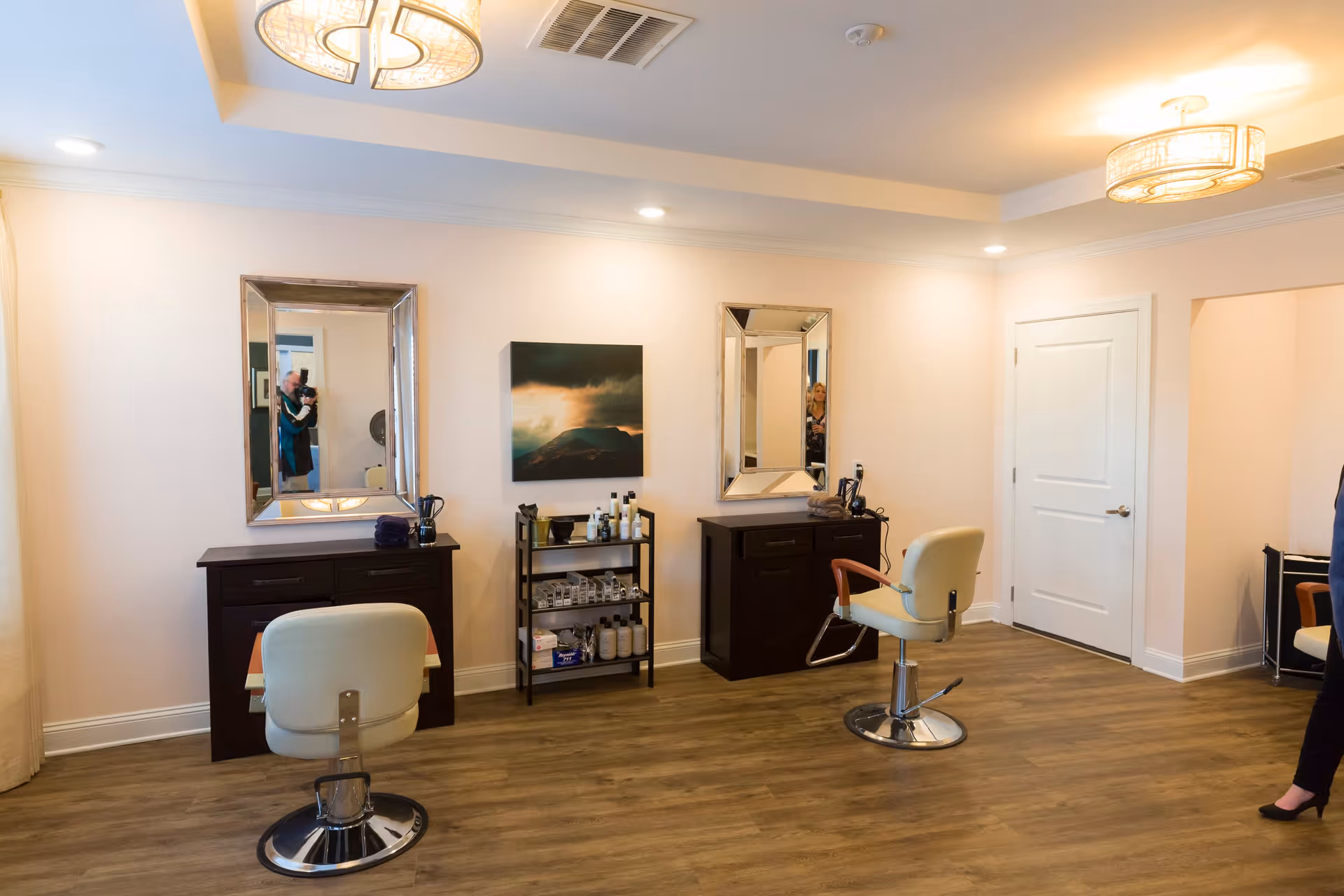 Interior view of a salon area with two styling stations, each with a large mirror, a dark wooden cabinet, and a cream-colored salon chair. There is a small shelving unit between the stations holding various hair care products. The room has wooden flooring, light-colored walls, and two ceiling light fixtures. A person is partially visible on the right side of the image.
