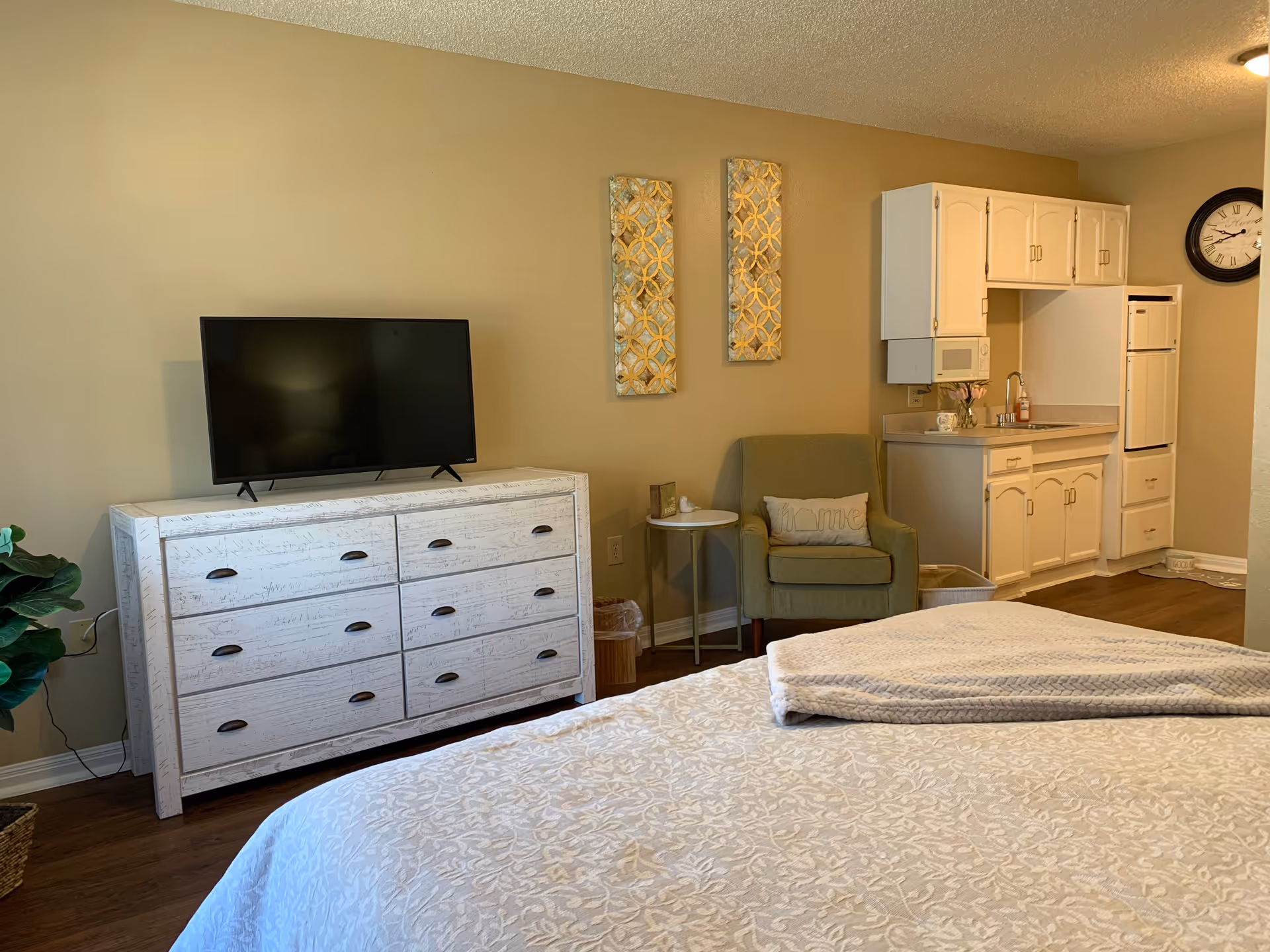 A cozy studio apartment featuring a bed with a patterned bedspread and folded blanket, a white dresser with a flat-screen TV on top, a green armchair with a decorative pillow, a small round side table, and a kitchenette with white cabinets, a microwave, a sink, and a refrigerator. The walls are beige with two decorative wall hangings and a clock.