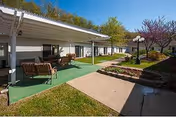 Outdoor patio area at a retirement center with covered seating including chairs and tables, adjacent to a building with white siding and large windows. There is a concrete walkway, green artificial turf, landscaped garden beds with shrubs and small trees, and a lamppost with multiple globes. Trees with spring foliage are visible in the background under a clear blue sky.