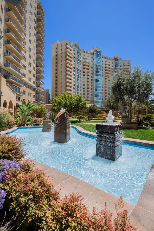 Outdoor courtyard area at Vi at La Jolla Village, featuring a clear blue sky, a water fountain with three stone pillars in a shallow pool, surrounded by landscaped greenery and flowers, with tall residential buildings in the background.
