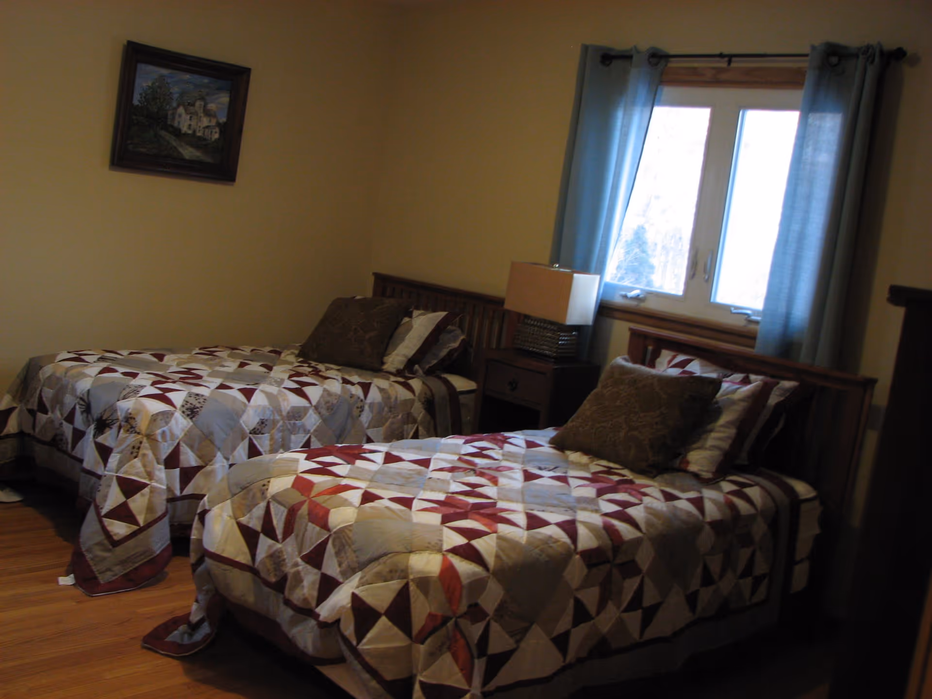 A bedroom with two twin beds covered in matching patchwork quilts in shades of red, white, and gray. Each bed has a pillow and a decorative cushion. Between the beds is a wooden nightstand with a lamp. A window with blue curtains is on the wall behind the nightstand, and a framed painting hangs on the adjacent wall. The room has wooden flooring and beige walls.