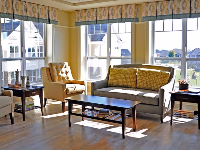 A bright living room area with large windows letting in natural light. The room features a gray sofa with two patterned yellow cushions, a beige armchair, a dark wooden coffee table, and two matching side tables. The windows have patterned valances, and outside views show neighboring buildings and greenery.