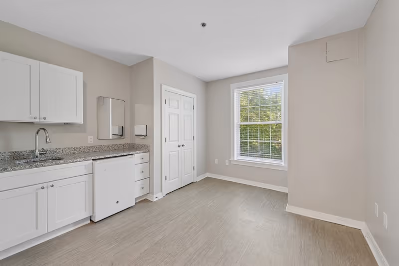 A clean, empty room with light beige walls and a large window with white blinds letting in natural light. On the left side, there is a small kitchenette area with white cabinets, a granite countertop, a sink, and a mini refrigerator. There is a double-door closet in the center of the back wall.