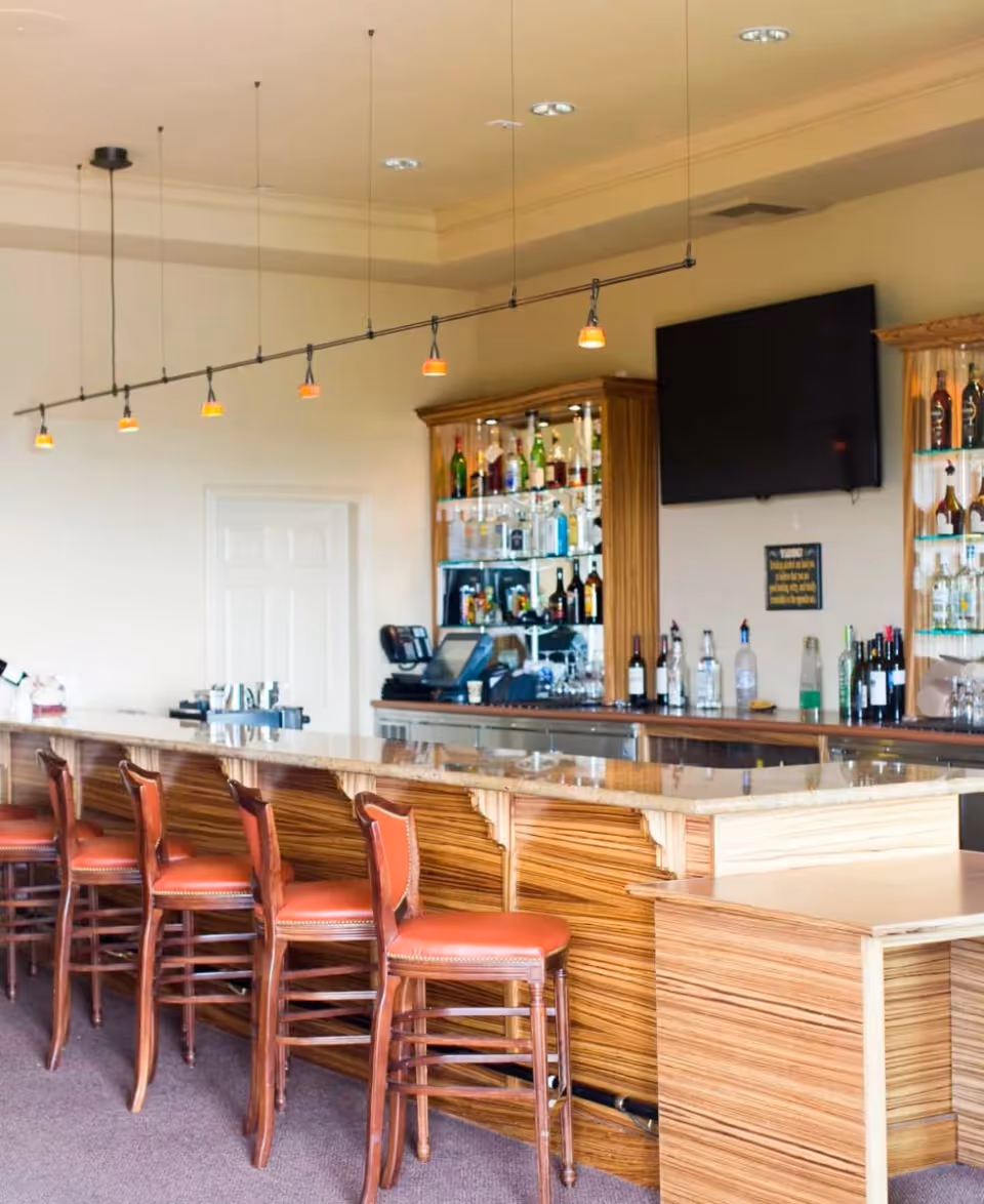 Interior view of a bar area with a long wooden counter and six high wooden chairs with red cushions. Behind the counter are shelves stocked with various bottles of liquor and a large flat-screen TV mounted on the wall. The ceiling has recessed lighting and a row of small pendant lights hanging above the bar.