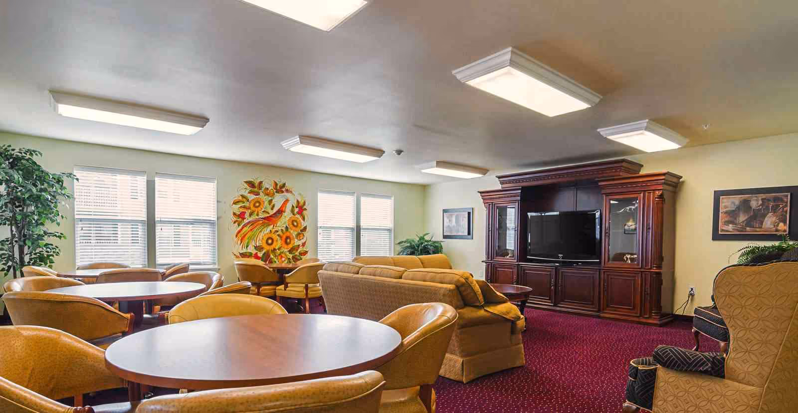 A spacious senior living common area with multiple round wooden tables surrounded by tan cushioned chairs. There is a large beige sofa facing a dark wooden entertainment center with a flat-screen TV. The room has large windows letting in natural light, a colorful wall mural of a bird and sunflowers, and framed artwork on the walls. The carpet is red with a subtle pattern, and there are several plants adding greenery to the space.