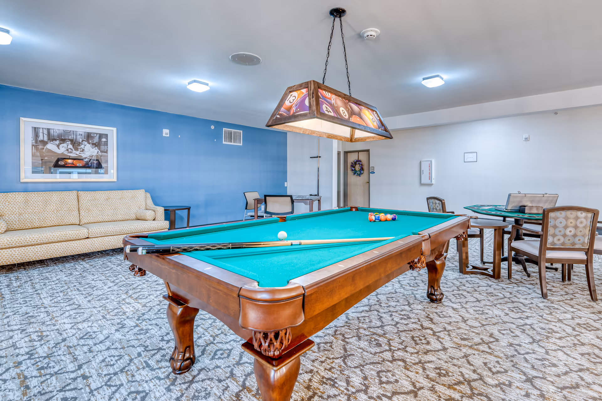 A recreational room with a pool table in the center, a hanging light fixture above it featuring billiard ball designs, a beige patterned sofa against a blue wall, a framed picture above the sofa, and a small table with chairs in the background.