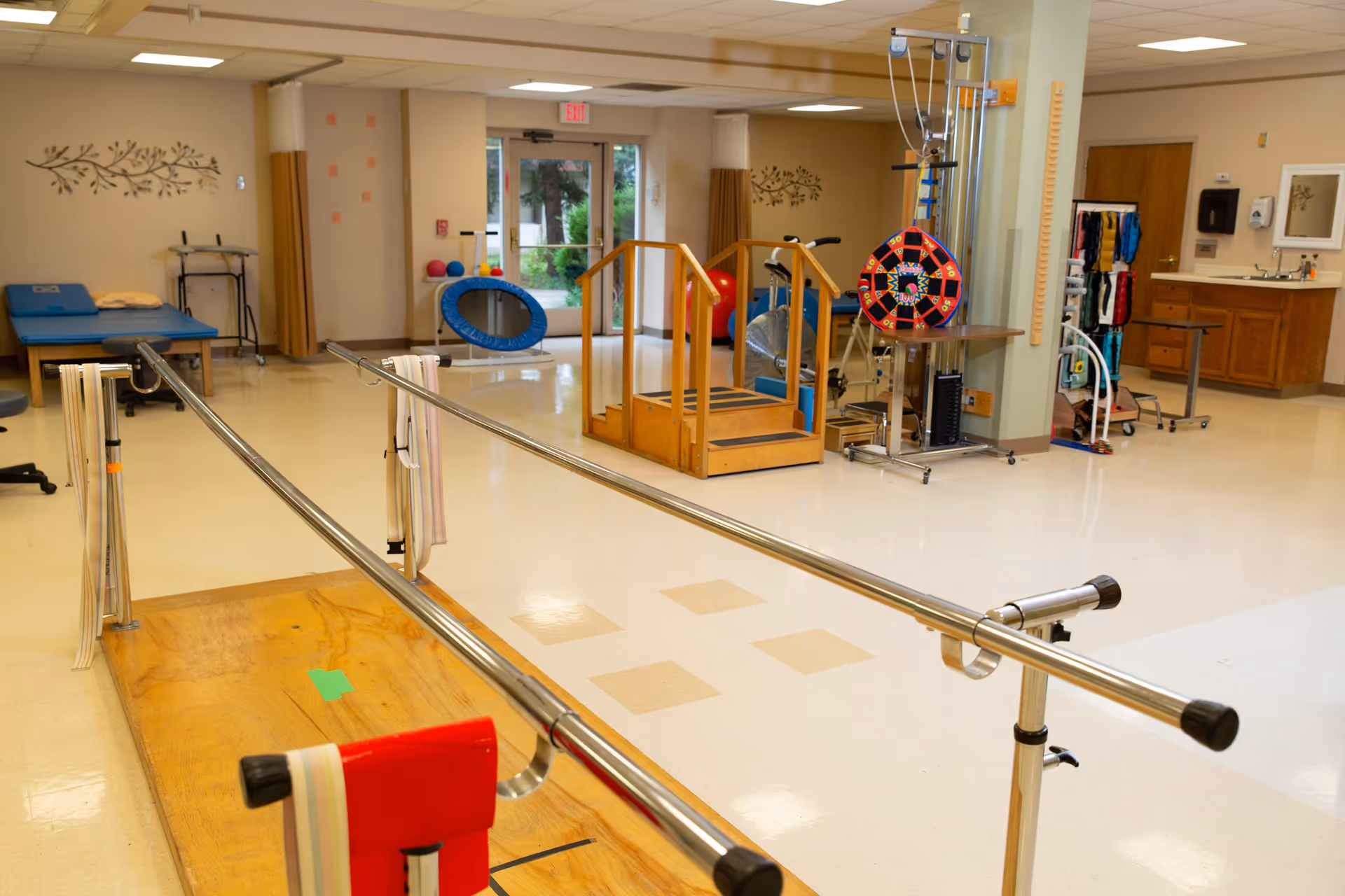 A physical therapy room with parallel bars for walking exercises, a wooden step platform, exercise balls, a blue therapy table, and various rehabilitation equipment. The room has beige walls, a tiled floor, and a window showing greenery outside.
