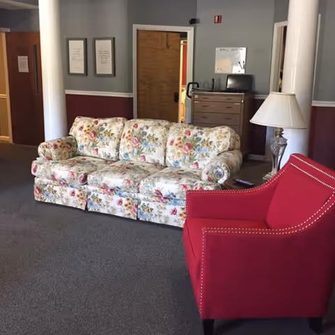 A cozy seating area in a living room with a floral patterned sofa and a red upholstered armchair. There is a side table with a lamp next to the armchair. In the background, there is a wooden dresser with a computer monitor on top and a whiteboard on the wall. The walls are painted gray with a maroon lower half and white trim.