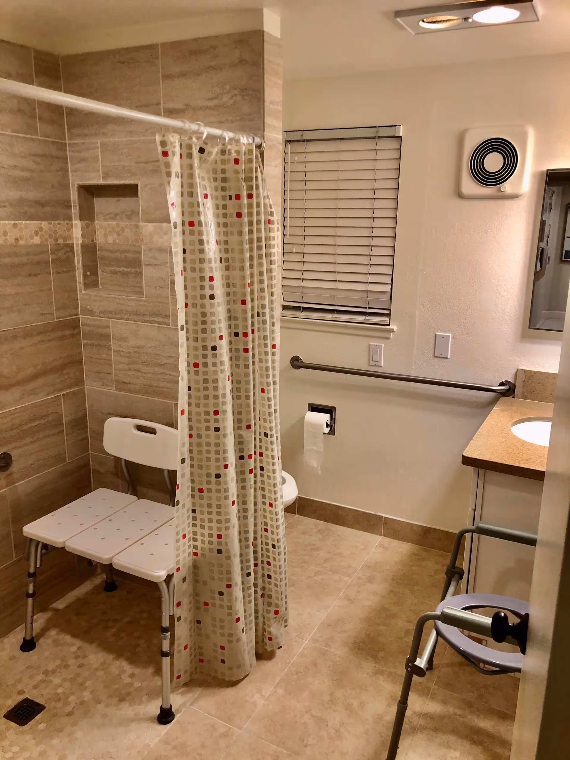 A bathroom with beige tiled walls and floor, featuring a walk-in shower area with a white shower chair and a patterned shower curtain. There is a window with closed blinds, a grab bar on the wall, a toilet paper holder, a sink with a countertop, and a walker near the door.