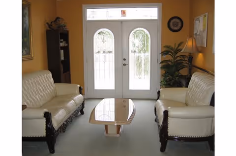 A cozy sitting area with two white leather sofas facing each other, a wooden coffee table in between, a tall plant with a lamp beside it, a bookshelf, and a decorative wall clock. The room has yellow walls and a glass-paneled double door letting in natural light.