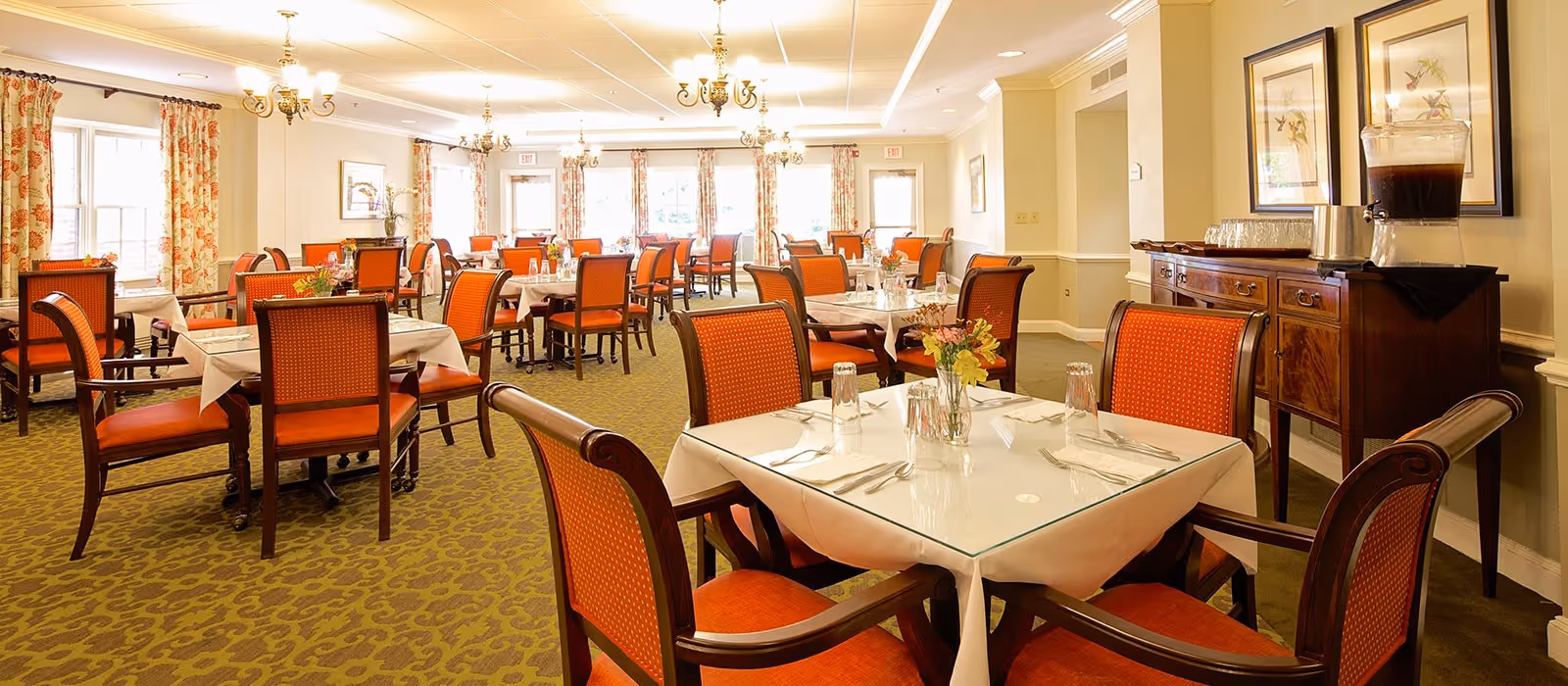 A bright dining room with multiple tables set with white linens and orange upholstered chairs, floral curtains, and a beverage station along the wall.