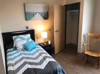 A small bedroom with a single bed featuring a chevron-patterned comforter in shades of gray, white, and teal. There are several pillows on the bed in matching colors. Next to the bed is a black nightstand with a lamp and a small device on it. The door to the room is open, revealing an empty closet. A wooden rocking chair is placed near the closet. A framed picture of a pier extending into water hangs on the wall above the bed.