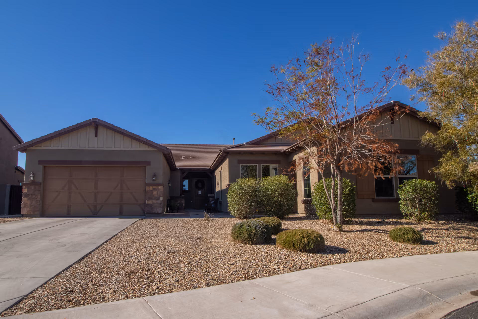 Front exterior view of a single-story residential building with a two-car garage, a driveway, and a landscaped front yard featuring small bushes and trees under a clear blue sky.