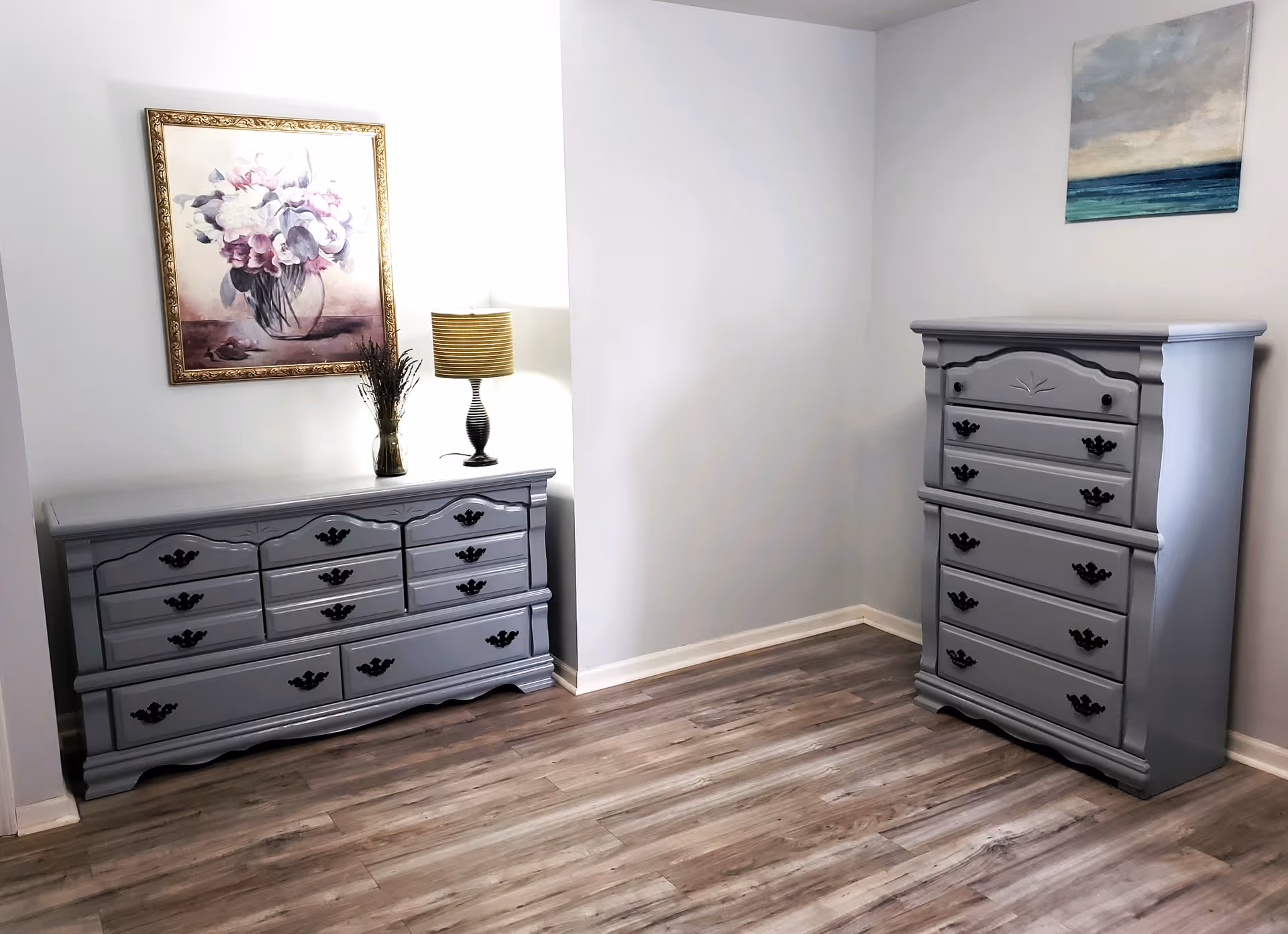 A light, sparsely furnished bedroom corner with two matching gray dressers, framed artwork, a table lamp, and wood-look flooring.