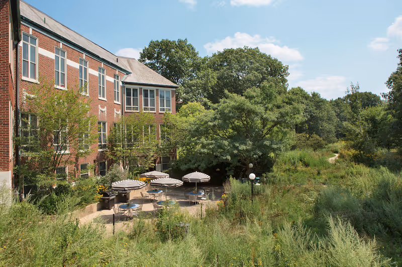 Brick assisted-living building overlooking a shaded outdoor patio with striped umbrellas and surrounding lush greenery.