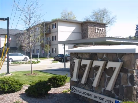 Exterior view of a multi-story residential building with the number 1777 displayed prominently on a stone sign in the foreground. The building has beige walls and multiple windows, with a few cars parked nearby and some small trees and bushes around the area.