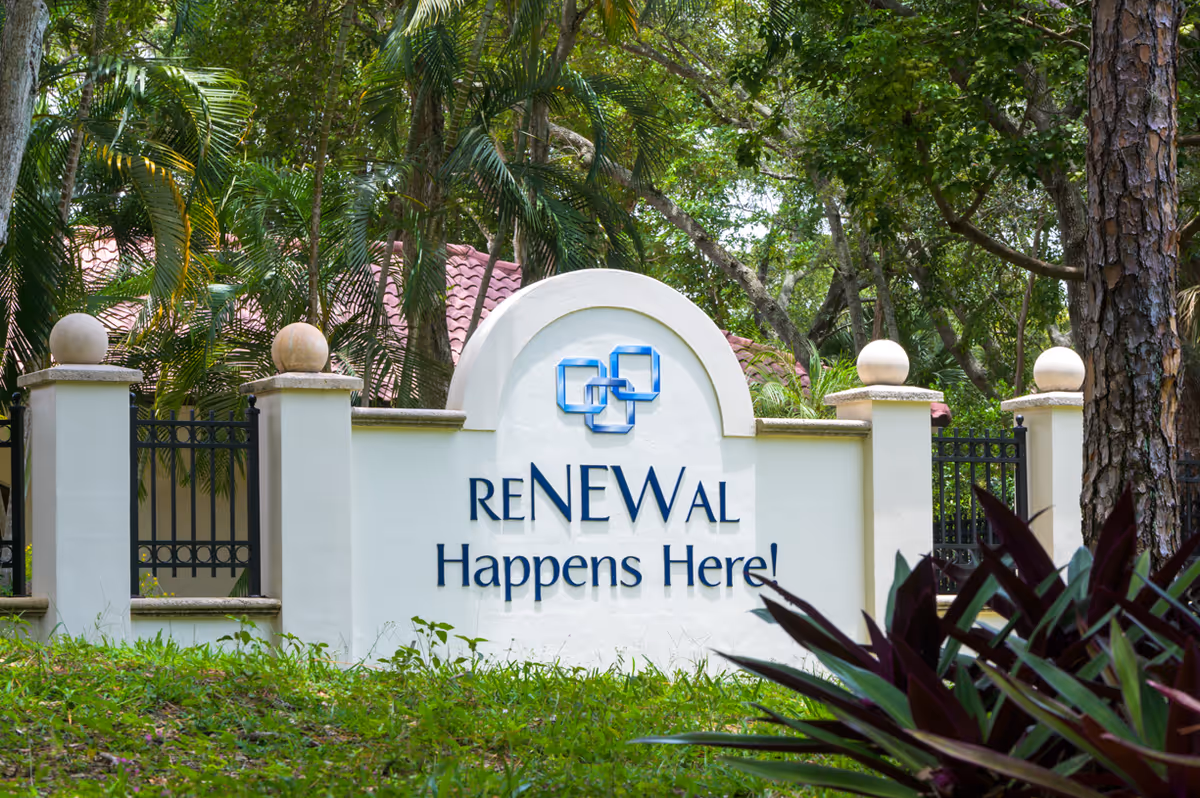 A white stone sign with decorative pillars and a blue logo that reads 'RENEWAL Happens Here!' surrounded by green trees and plants.